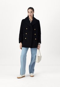Mujer con el cabello trenzado que lleva un abrigo negro de doble botón, jeans de color azul claro, zapatillas blancas y sostiene un pequeño bolso blanco.