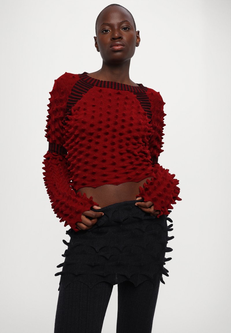 Roter strukturiert geschnittener Pullover mit erhobenen Punktmuster und schwarzen horizontalen Streifen am Ausschnitt, kombiniert mit einem schwarzen geschichteten Minirock.