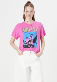 Rosa bomull T-skjorte med grafisk trykk av et band, med grønn tekst og blå bakgrunn. Kombinert med hvite denimshorts.