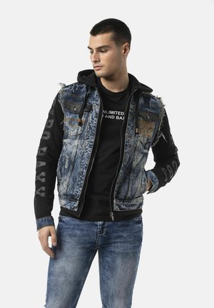 Junger Mann, der einen schwarzen Hoodie mit ärmelloser Jeansjacke und blauen Jeans trägt, schaut zur Seite mit einer Hand in der Tasche.