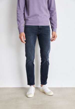 Person trägt eine dunkelblaue Slim-Fit-Jeans, weiße Sneaker und ein langärmeliges lavendelfarbenes Oberteil und steht an einer weißen Wand.