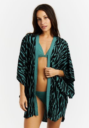 Vrouw in teal bikini en bijpassend teal en zwart zebraprint kimono, staand tegen een witte achtergrond.