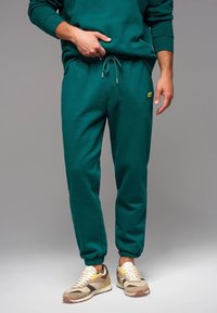 Los pantalones de chándal verdes cuentan con una cintura con cordón, puños elásticos y un bolsillo lateral, combinados con zapatillas beige y amarillas. Textura suave.