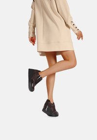 Sneaker neri realizzati in un mix di materiali testurizzati con una finitura lucida e lacci, abbinati a un vestito oversize beige.