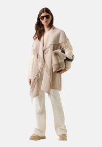 Béžový šál s třásněmi, přehozený přes krémový top, bílé kalhoty a béžové tenisky. Modelka drží huňatou tašku a nosí oversized sluneční brýle.