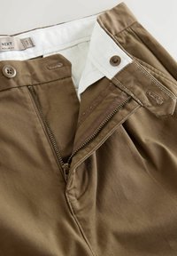 Dark brown trousers with a zip closure, featuring a white inner waistband and brown button accents. Fabric has a smooth, woven texture.