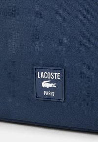 Navy blå stoffveske med teksturert overflate, med et rektangulært Lacoste-logo i hvitt og navy. Minimalistisk design med tydelig merkevare.