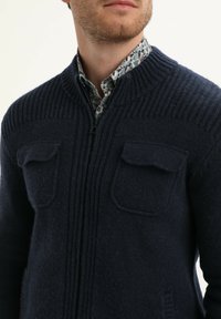 Cardigan in maglia navy con zip sul davanti, colletto a coste e due tasche sul petto. Il tessuto ha una consistenza morbida e un motivo sottile sotto.