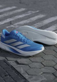 Paire de chaussures de course bleues avec semelles blanches et rayures grises, placées sur un pavé hexagonal près d’un passage piéton.