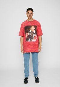 Red tie-dye oversized t-shirt featuring a central graphic print. Paired with light blue jeans and black sneakers. Neutral backdrop.
