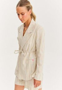 Pruhovaný blazer vyrobený z lehkého materiálu, s pastelovými vertikálními pruhy, šňůrkovým páskem a růžovým logem na nášivce poblíž kapsy.