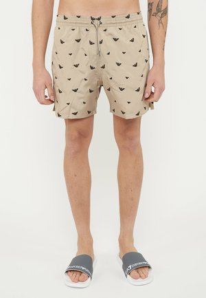 Beige Shorts mit schwarzen, wiederholten Logos, getragen von einer Person mit Tätowierungen an einem Arm, kombiniert mit grauen Slidesandalen mit weißen Sohlen.
