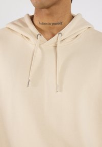 Beige hoodie with a soft texture, adjustable drawstrings, silver hardware, and a lined hood. Minimalist design, no visible patterns.