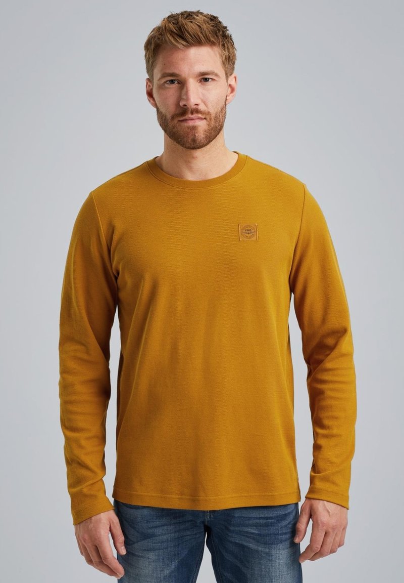 Langärmliges senfgelbes T-Shirt aus strukturiertem Stoff mit Rundhalsausschnitt und kleinem Logoschild auf der linken Brust. Kombination mit blauen Jeans.