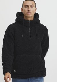 Svart fleece hettegenser med kvart glidelås, hette og frontkangaroo-lomme. Strukturert, plysj stoff med justerbare snor for hetten.