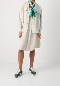 Camisa de lino beige con botones, vestido beige a juego, zapatillas Nike verdes y blancas, y una bufanda de patrón verde azulado atada al cuello.