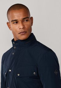 Mand med kort hår iført en marineblå zip-up jakke med trykknaplommer og høj krave, der kigger til venstre imod en enkel baggrund.