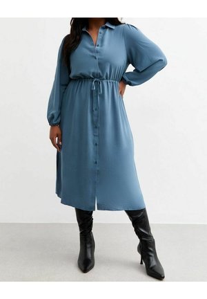 Femme portant une robe midi bleue à manches longues boutonnée avec une ceinture à la taille et des bottines noires à talons pointus, debout, une main sur la hanche.