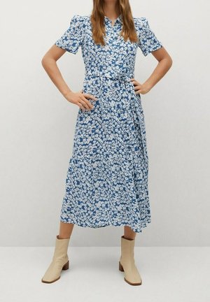 Frau trägt ein blaues Midikleid mit weißem Blumenmuster, kurzen Puffärmeln, einem Taillengürtel und beigen Ankle-Boots, steht mit den Händen in der Hüfte.