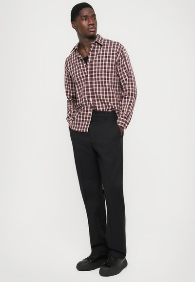 Uomo che indossa pantaloni neri, sneakers nere e una camicia a scacchi bordeaux e bianca, con le mani in tasca, che guarda a sinistra su uno sfondo neutro.
