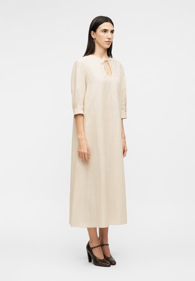 Γυναίκα με μπεζ φόρεμα μακρυμάνικο μέχρι τον αστράγαλο με λαιμόκοψη σε σχήμα κλειδαρότρυπας, φορά καφέ ψηλοτάκουνα παπούτσια Mary Jane, στέκεται μπροστά σε λευκό φόντο.