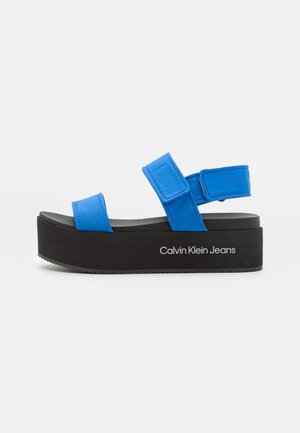 Blue platform sandals with black outsole, featuring two adjustable straps and "Calvin Klein Jeans" printed on the side. Soft material.
