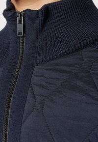 Veste zippée bleu marine foncé avec un col montant côtelé et un panneau matelassé texturé, dotée d'une fermeture éclair noire et d'une finition lisse.