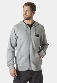 Sudadera gris con cremallera en tejido suave, con un bolsillo frontal tipo canguro, capucha ajustable y logo de Helly Hansen en la parte izquierda del pecho.