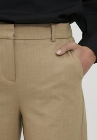Beige maßgeschneiderte Hose mit glatter Textur, seitlichen Taschen und flacher Vorderseite. Der Bund verfügt über einen Gürtelschlaufen und eine ordentliche Verarbeitung.