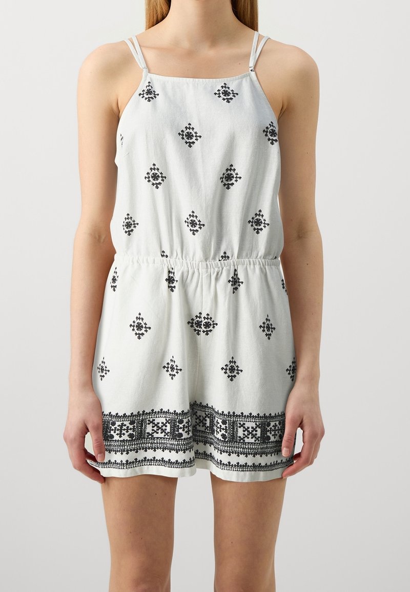 Femme portant une combinaison blanche sans manches avec de fines bretelles et des motifs géométriques noirs sur le tissu.