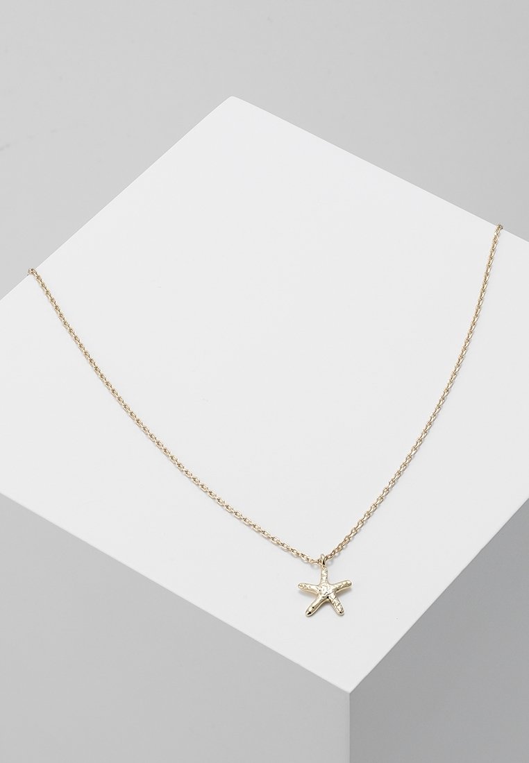 Collier fin en chaîne d’or avec un petit pendentif en forme d’étoile de mer reposant sur une surface blanche inclinée.