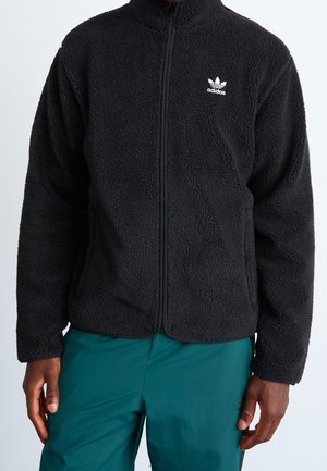 Mann trägt eine schwarze Adidas Fleecejacke mit durchgehendem Reißverschluss und türkisfarbene Hose vor einfachem hellem Hintergrund.