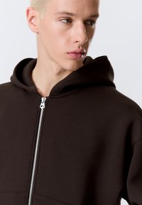 Junge Person mit hellblonden Haaren, die einen dunkelbraunen Kapuzenpullover mit Reißverschluss trägt und nach rechts blickt, vor einem einfarbigen hellen Hintergrund.