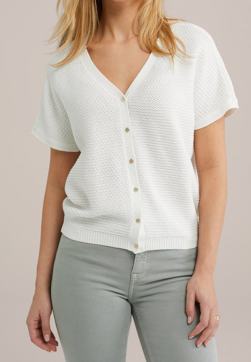 Žena v bílém krátkém pleteném cardigan s knoflíky a světle šedých přiléhavých kalhotách, stojící na jednoduchém pozadí.