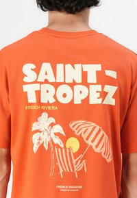 Oranje katoenen T-shirt met een grafische print van palmbomen, zon en strandstoel, met de tekst "SAINT-TROPEZ" in dikke crèmelettertype.