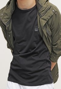 Giacca leggera verde oliva con zip sopra una maglietta nera a maniche corte, con etichetta rettangolare cucita sulla maglietta.