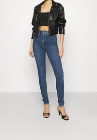 Chaqueta corta de cuero sintético negra, top de corte ajustado negro y jeans ajustados azul de cintura alta. La modelo lleva sandalias negras de tacón abierto.