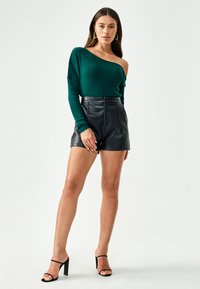 Maglione verde a coste con spalle scoperte, pantaloni shorts neri in pelle a vita alta, sandali neri con tacco alto e cinturini. Texture liscia e design aderente.