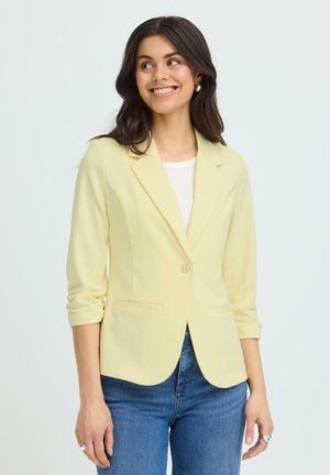 Jeune femme souriante, portant un blazer jaune clair aux manches retroussées, un haut blanc et un jean bleu, sur un fond clair.