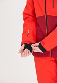 Rote und dunkelrote Jacke mit hohem Kragen, ausgestattet mit einem Klettverschluss verstellbaren Bündchen und Logodetail am Ärmel. Glatte Stoffstruktur.