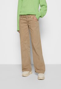 Calças largas bege em mistura de algodão com bolsos, fecho de botão à frente e um corte reto. Combinadas com um suéter verde e sapatos claros.