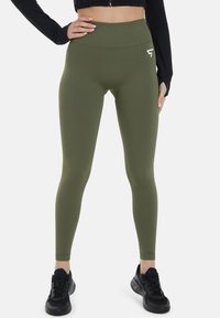 Oliivinvihreät legginsit korkealla vyötäröllä, valmistettu joustavasta materiaalista, jossa on pieni valkoinen logo. Mustat lenkkarit ovat käytössä.
