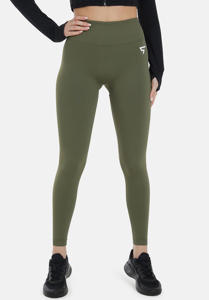 Oliivinvihreät legginsit korkealla vyötäröllä, valmistettu joustavasta materiaalista, jossa on pieni valkoinen logo. Mustat lenkkarit ovat käytössä.