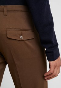 Gros plan d'une personne portant un pantalon marron avec une poche arrière boutonnée et un haut à manches longues bleu marine, main reposant sur le côté.