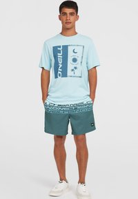 Lyseblå t-shirt med grafisk print af palmer og sol; parret med mørk teal shorts med hvid logo-elastik og lap.