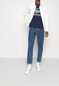 Muž oblečený v bílé džínové bundě přes námořnické tričko s logem, modrých džínách, bílých ponožkách a bílých teniskách stojící na šedém pozadí.