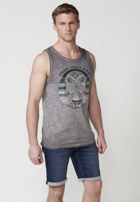 Siva brezrokavnik s potiskom lobanje, ki vsebuje zapletene oblikovalske elemente. V kombinaciji s temnimi denim kratkimi hlačami, zavitimi na robu.