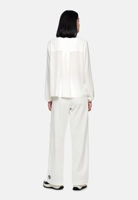 Weiße Bluse mit langen Ärmeln und transparentem Oberteil am Rücken, kombiniert mit lockeren weißen Hosen. Der Stoff wirkt weich mit glatter Textur. Schwarzes Logo auf der Hose.