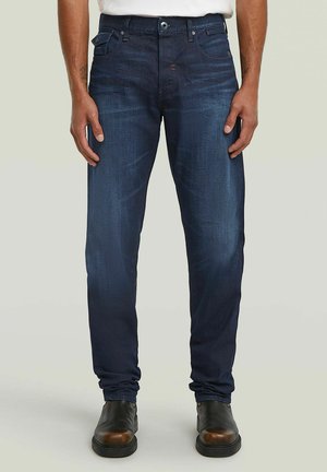 Mann steht, trägt dunkelblaue Slim-Fit-Jeans und schwarze Lederschuhe vor einem einfarbigen hellen Hintergrund.