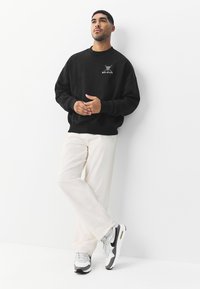 Sort sweatshirt med fedt grafisk motiv på brystet, kombineret med lyse, løstsiddende bukser og grå Nike-sneakers med detaljer.